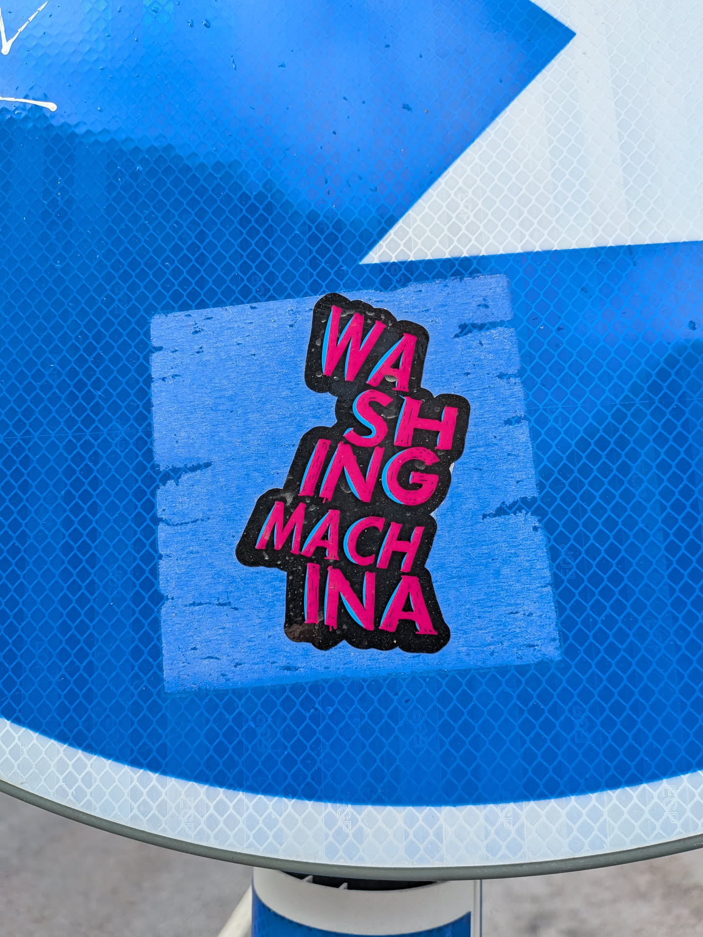The sticker is roughly rectangular with an irregular, slightly wavy outline that follows the stacked text. It is applied to a reflective blue surface with a diamond grid pattern. The sticker's background is a faded light blue. The main content is the phrase "WASHING MACHINE" stacked vertically. The letters are bold, blocky, and feature a striking neon pink/magenta fill with a bright cyan/light blue drop shadow, all outlined in thick black. The overall aesthetic is vibrant and retro. There are visible signs of wear, including slight peeling at the edges and some dirt or grime.