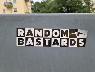 Street sticker A black and white sticker with the words "RANDOM BASTARDS" arranged in a checkered pattern.  A small graphic of two birds is included in the design. The sticker is affixed to a grey surface.