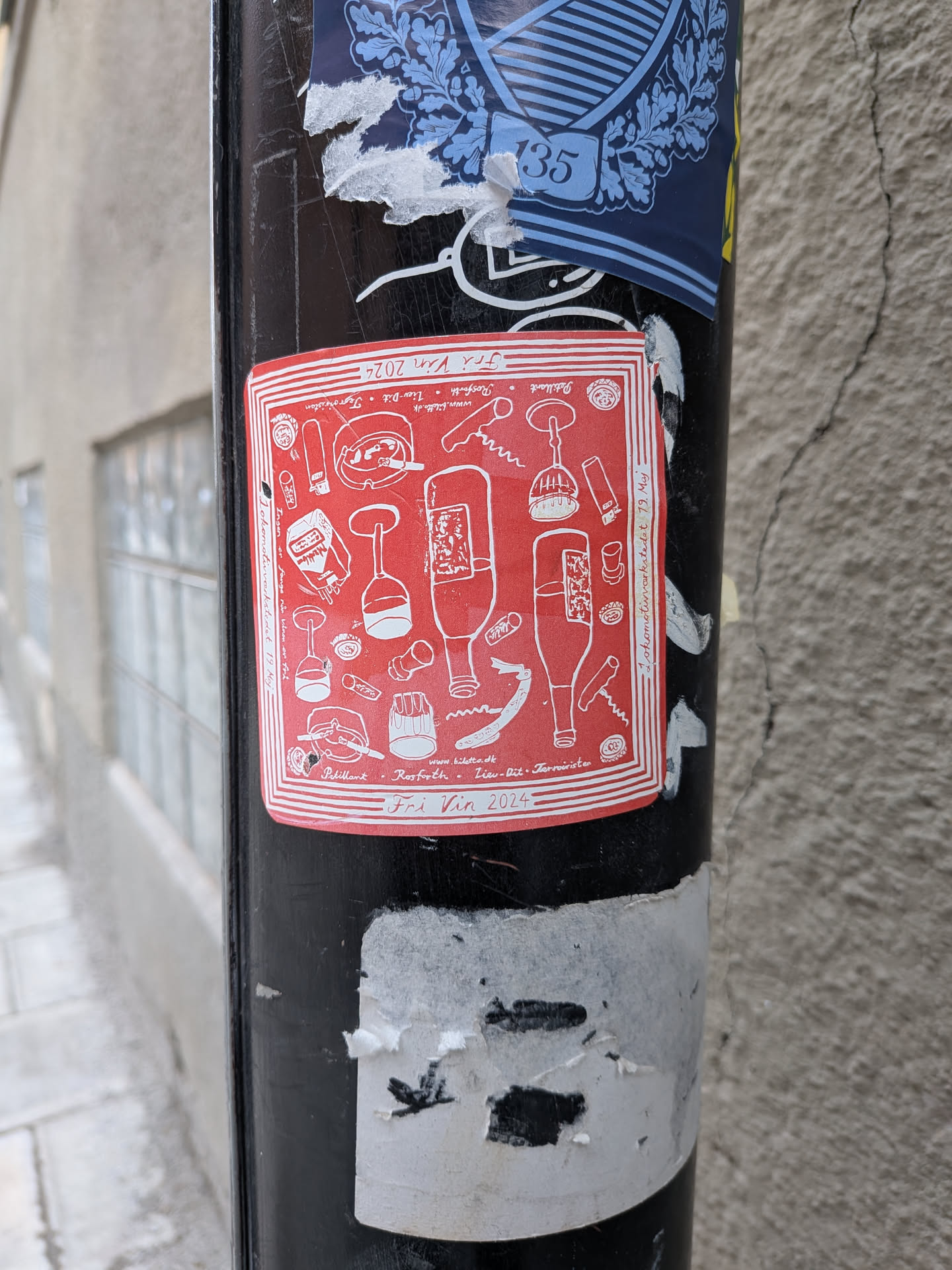 A square red sticker with white line art illustrations, depicting various wine-related items such as wine bottles, wine glasses, corkscrews, bottle openers, and corks. The illustrations are arranged in a whimsical, scattered pattern. Text is present around the border and within the design, including event details and names. The sticker has a thin white border with multiple parallel lines, giving it a framed appearance. It is affixed to a black pole, showing some minor wear and tear.