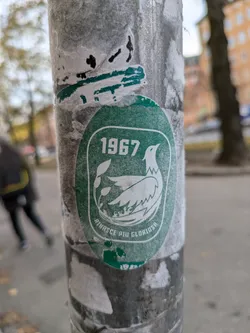Street sticker A green oval sticker with the year 1967 and the text "Rinase Più Gloriosa" which translates from Italian to "Reborn More Glorious."  The sticker depicts a stylized bird surrounded by floral elements. The sticker is affixed to a weathered pole, showing some wear and tear.