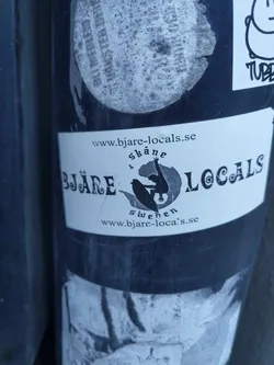 Street sticker A black and white sticker featuring the Bjäre Locals logo, which includes a silhouette of a person surfing or doing a similar activity, with the text 'Bjäre Locals' and website address www.bjare-locals.se.  The sticker is adhered to a dark surface and appears slightly worn.