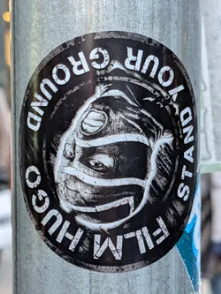A circular sticker with a black background and white text and illustration. The central illustration is a black and white sketch of a face wearing a mask or helmet, with visible eyes peering out. The text "STAND YOUR GROUND" is written in a circular pattern around the top half of the sticker in bold white capital letters. The text "HUGO FILM GO" is written in a circular pattern around the bottom half of the sticker in bold white capital letters, with "GO" appearing to be slightly cut off or obscured. The overall style is graphic and somewhat gritty.