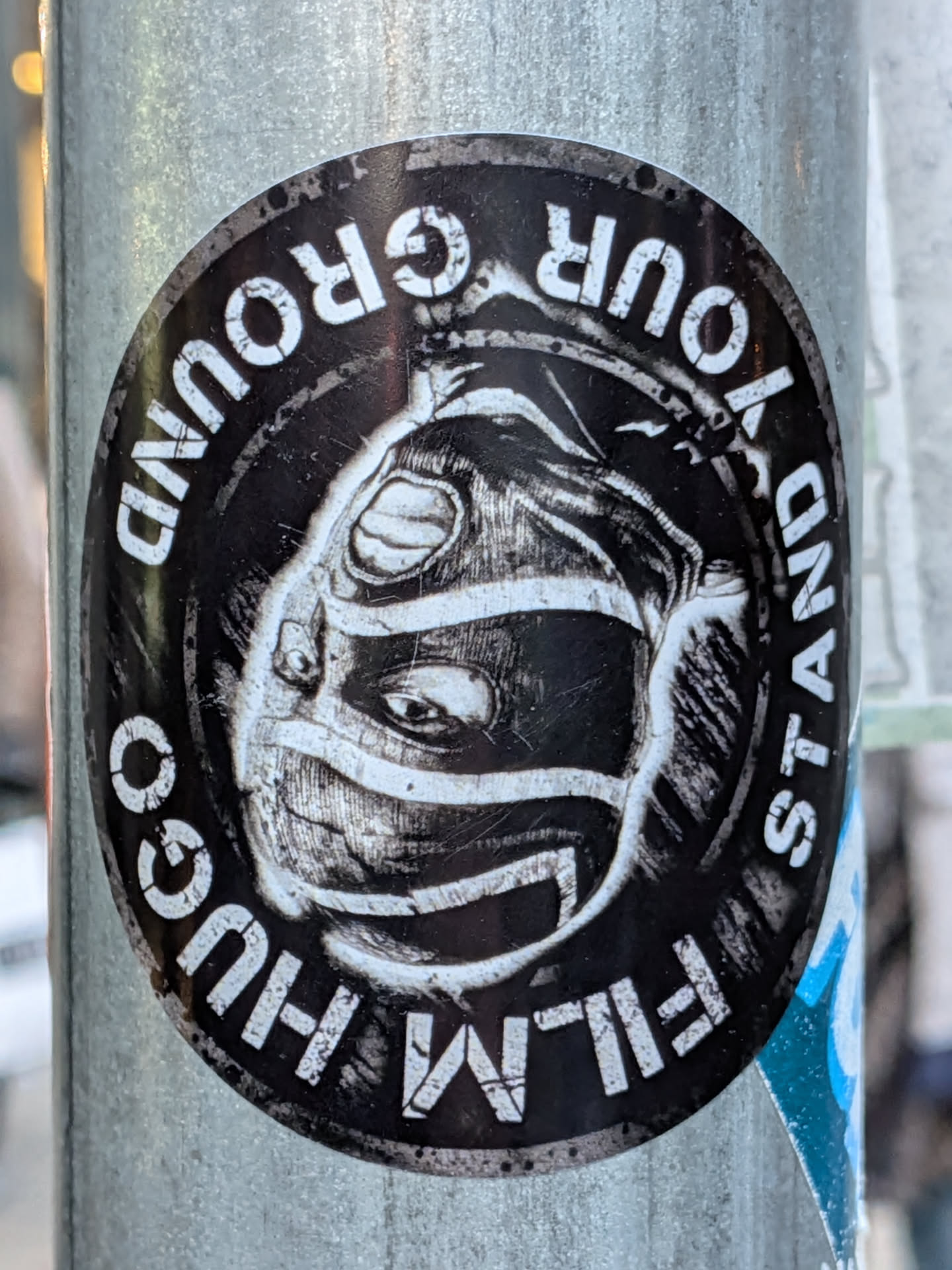 A circular sticker with a black background and white text and illustration. The central illustration is a black and white sketch of a face wearing a mask or helmet, with visible eyes peering out. The text "STAND YOUR GROUND" is written in a circular pattern around the top half of the sticker in bold white capital letters. The text "HUGO FILM GO" is written in a circular pattern around the bottom half of the sticker in bold white capital letters, with "GO" appearing to be slightly cut off or obscured. The overall style is graphic and somewhat gritty.