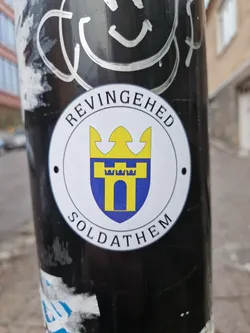 Street sticker Close-up of a circular sticker displaying the coat of arms of Soldathem, a locality in Revingehed, Sweden. The emblem showcases a yellow and blue shield with a castle-like structure and a crown. The text 'REVINGEHED' and 'SOLDATHEM' surrounds the emblem.