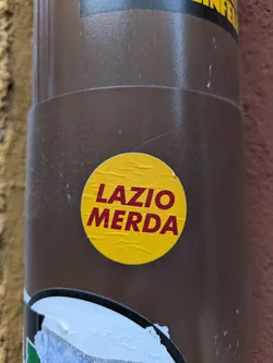 Street sticker A round yellow sticker with the words "Lazio Merda" written in maroon. The sticker is affixed to a brown cylindrical object.