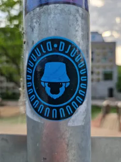 Street sticker A round sticker with a black background and bright blue accents. It features a cartoon illustration of a bearded man wearing a bucket hat. The text "DJUU" is written in a circle around the main image. The sticker appears to be affixed to a weathered metal pole outdoors.