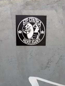 Street sticker A square black sticker with a white circular emblem. The emblem depicts a character in a balaclava alongside a spray paint can, flanked by two stylized chestnut leaves. Cyrillic text is arched at the top, and 'HOOLIGANS' is written at the bottom.