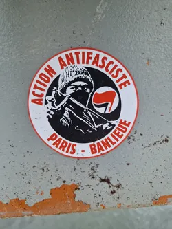 Street sticker A round sticker with a black and white illustration of a hooded person holding a slingshot. The text around the illustration says "Action Antifasciste Paris-Banlieue" in French, meaning "Antifascist Action Paris-Suburbs".  A red flag symbol is also present. The sticker appears to be placed on a weathered surface.