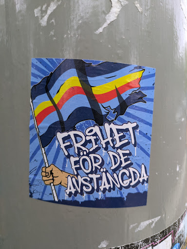 This sticker shows a hand holding a flag with horizontal stripes of blue, yellow, and red. The background is blue with sunburst patterns. The text "FRIHET FÖR DE AVSTÄNGDA" is written in a graffiti style.