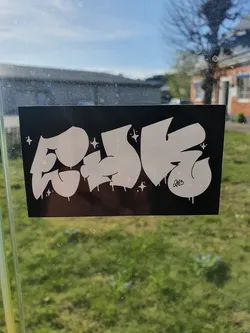 This sticker features a stylized graffiti-like design with the word "FOJIE" in bold, white letters on a black background. The letters have a glossy, almost dripping effect, and there are small star details scattered around. The artist's signature is subtly placed at the bottom right corner. 