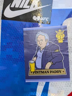 Street sticker A sticker featuring an illustration of a man, possibly named Paddy, holding a pint of beer. The style is cartoonish and features a man with a slight beer belly wearing a purple jacket and light blue shirt.  He is standing next to a small sign which reads 'Pintman Paddy'.  Another smaller graphic of a crowned head appears in a corner of the sticker.