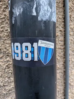 Street sticker A rectangular sticker with a dark blue background, adhered to a black pole. On the left side, the year '1981' is prominently displayed in large, silver-outlined numbers, filled with a light blue and dark blue checkered pattern. To the right, there is a shield-shaped emblem. The top section of the shield is white, featuring the text 'Järnkammerna' in a decorative script font, with 'stockholm' in a smaller, sans-serif font directly below it. The main body of the shield is vertically striped in alternating shades of blue and white. The sticker shows some minor scuffs and wear, indicating its outdoor placement.