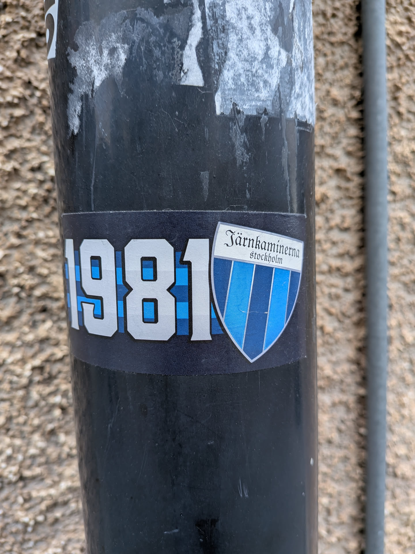 A rectangular sticker with a dark blue background, adhered to a black pole. On the left side, the year '1981' is prominently displayed in large, silver-outlined numbers, filled with a light blue and dark blue checkered pattern. To the right, there is a shield-shaped emblem. The top section of the shield is white, featuring the text 'Järnkammerna' in a decorative script font, with 'stockholm' in a smaller, sans-serif font directly below it. The main body of the shield is vertically striped in alternating shades of blue and white. The sticker shows some minor scuffs and wear, indicating its outdoor placement.