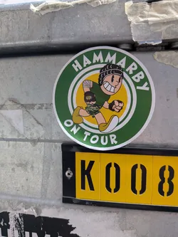 Street sticker Circular sticker with a green border reading 'HAMMARBY ON TOUR'. The center features a yellow background with a retro cartoon character running, wearing a green shirt, camo shorts, and a black head guard.
