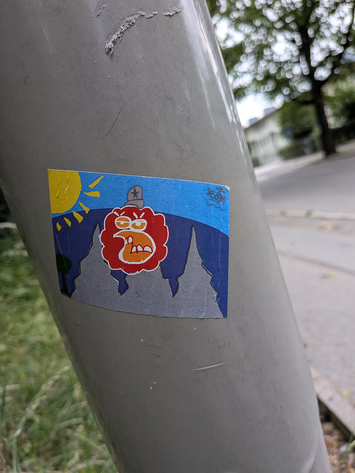 A rectangular sticker depicting an angry red character with a star-topped hat. The background features grey mountain peaks, dark blue hills, a yellow sun, and a blue sky. A small signature '2024 KOSM' is in the top right corner.