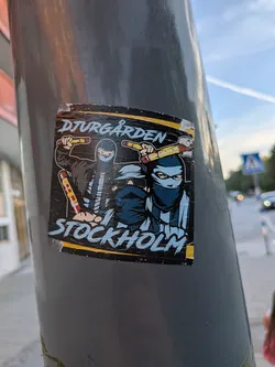Street sticker The sticker features two figures wearing masks and dark clothing, holding what appear to be flares or batons. The top text reads 'DJURGÅRDEN' in a stylized, distressed font with a yellow outline. Below the figures, the text 'STOCKHOLM' is written in a similar, bold, blue font with white highlights. The overall color scheme is dark with accents of yellow, blue, and red from the flares. The sticker has a slightly worn appearance, suggesting it has been exposed to the elements.