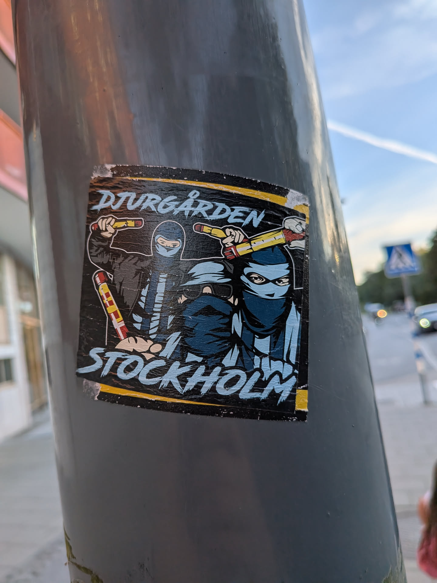 The sticker features two figures wearing masks and dark clothing, holding what appear to be flares or batons. The top text reads 'DJURGÅRDEN' in a stylized, distressed font with a yellow outline. Below the figures, the text 'STOCKHOLM' is written in a similar, bold, blue font with white highlights. The overall color scheme is dark with accents of yellow, blue, and red from the flares. The sticker has a slightly worn appearance, suggesting it has been exposed to the elements.