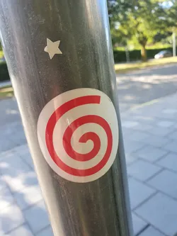 A round sticker with a red spiral design on a white background.  It's affixed to a gray metal pole outdoors. There is also a small white star sticker above it.