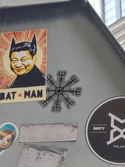 Street sticker This sticker depicts a Vegvisir, a symbol from Icelandic magic, said to guide travelers.  It's a circular symbol with eight arms pointing outwards, each with a different pattern. The sticker has a simple, minimalist design, with the Vegvisir in grey on a white background. It is placed amongst other stickers on a grey surface.