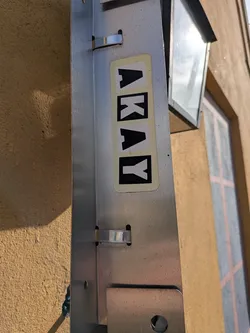 Street sticker A rectangular sticker with rounded corners and a pale yellow background is affixed vertically to a metallic fixture. The sticker features the word 'AKAY' spelled out in individual square blocks. The first three letters ('A', 'K', 'A') are black on white squares, while the final letter ('Y') is white on a black square.
