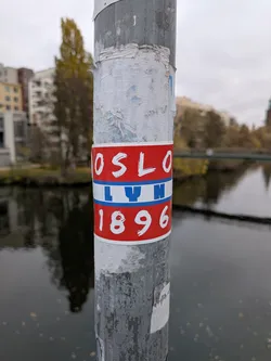 Street sticker The sticker is circular and features three horizontal bands. The top and bottom bands are red with white text, while the middle band is blue with white text. The top band reads 'OSLO' in a stylized white font. The middle band reads 'LYN' in a similar white font. The bottom band reads '1896' in white numerals. The sticker is affixed to a metal pole, with some peeling and wear visible on the pole's surface.