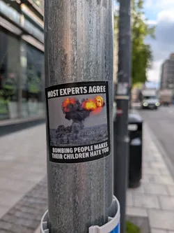 Street sticker A rectangular sticker with a black border, featuring a powerful anti-war message. The top section displays the text 'MOST EXPERTS AGREE' in bold white capital letters against a black background. The central image depicts a large, dark grey mushroom cloud explosion with intense orange and yellow flames at its peak, rising above a muted, grey-toned city skyline. The bottom section contains the text 'BOMBING PEOPLE MAKES THEIR CHILDREN HATE YOU' in bold white capital letters, also against a black background. The overall aesthetic is somber and impactful, using a limited color palette of black, white, grey, and fiery orange/yellow.