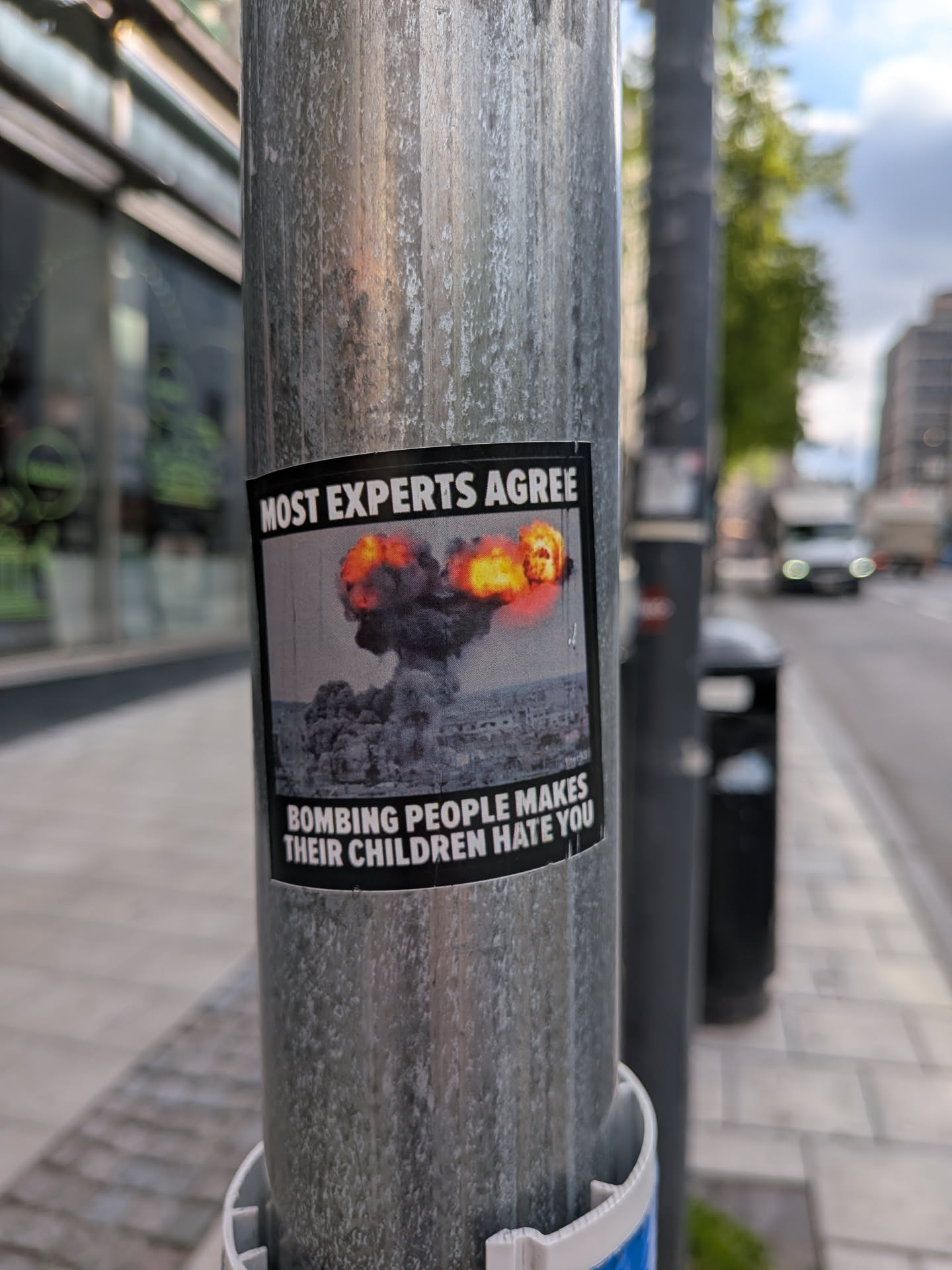 A rectangular sticker with a black border, featuring a powerful anti-war message. The top section displays the text 'MOST EXPERTS AGREE' in bold white capital letters against a black background. The central image depicts a large, dark grey mushroom cloud explosion with intense orange and yellow flames at its peak, rising above a muted, grey-toned city skyline. The bottom section contains the text 'BOMBING PEOPLE MAKES THEIR CHILDREN HATE YOU' in bold white capital letters, also against a black background. The overall aesthetic is somber and impactful, using a limited color palette of black, white, grey, and fiery orange/yellow.