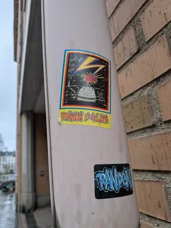 Street sticker The central sticker is rectangular with a colorful, slightly worn border featuring blue, red, and yellow stripes. The main artwork is set against a dark, textured background, possibly representing a night sky or a gritty surface. A prominent yellow lightning bolt with a red shadow strikes from the upper left towards the center. Below it, a white, detailed illustration of a capitol building dome is visible. Directly above the dome, a red and yellow explosion or burst of light emanates, accompanied by small blue dots suggesting sparks. Below the main image, a bright yellow banner contains the text 'BRÄNN SALVA' in bold, red, somewhat jagged letters. The sticker shows signs of wear and slight peeling at the edges, indicating it is a physical sticker applied to a surface.