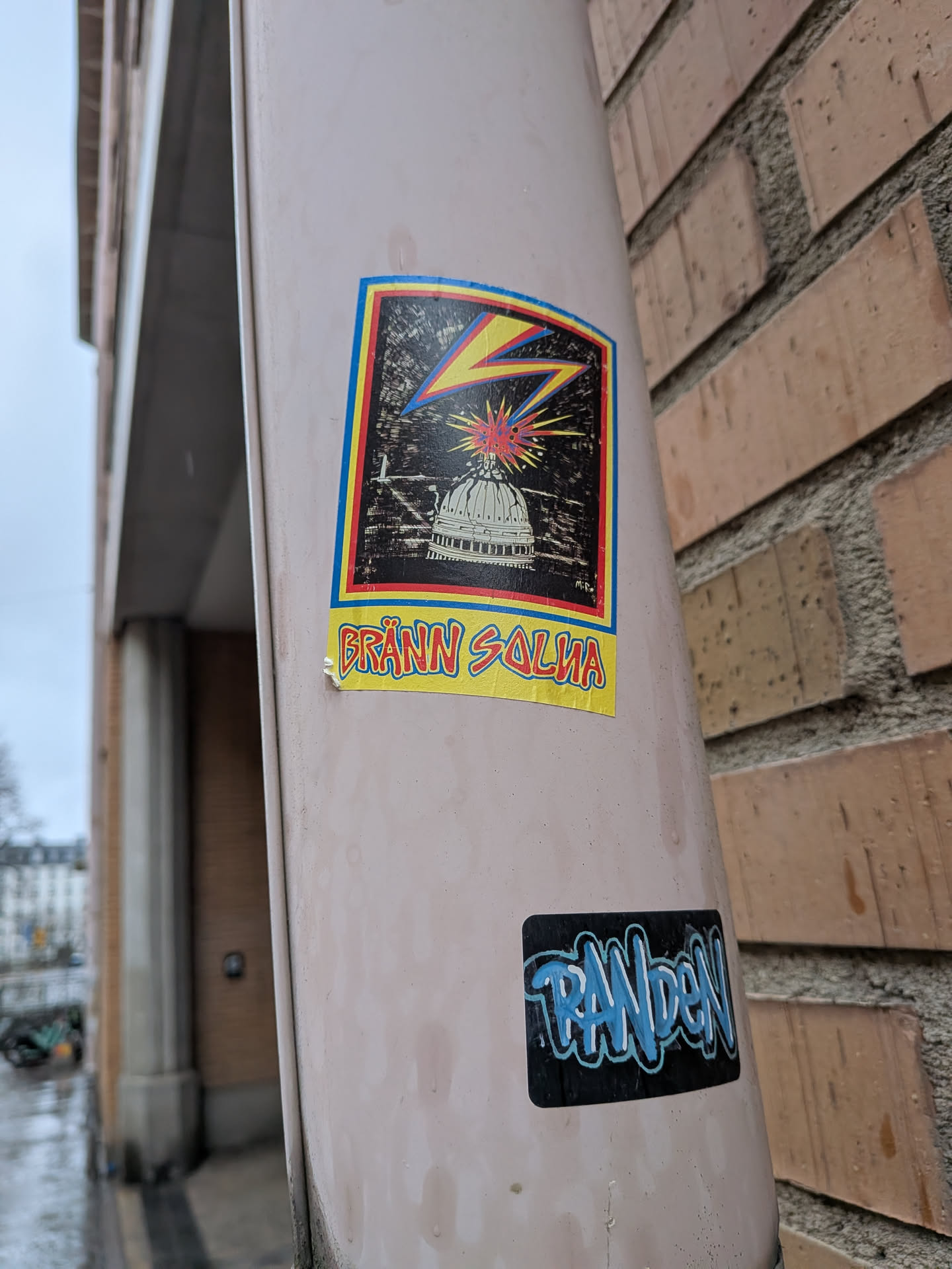 The central sticker is rectangular with a colorful, slightly worn border featuring blue, red, and yellow stripes. The main artwork is set against a dark, textured background, possibly representing a night sky or a gritty surface. A prominent yellow lightning bolt with a red shadow strikes from the upper left towards the center. Below it, a white, detailed illustration of a capitol building dome is visible. Directly above the dome, a red and yellow explosion or burst of light emanates, accompanied by small blue dots suggesting sparks. Below the main image, a bright yellow banner contains the text 'BRÄNN SALVA' in bold, red, somewhat jagged letters. The sticker shows signs of wear and slight peeling at the edges, indicating it is a physical sticker applied to a surface.