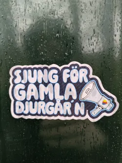 Street sticker The sticker features the text 'Sjung för Gamla Djurgår'n', which translates to 'Sing for Old Djurgården' in English. It also includes a stylized illustration of a megaphone with a color accent. The sticker appears to be affixed to a dark green surface with raindrops.