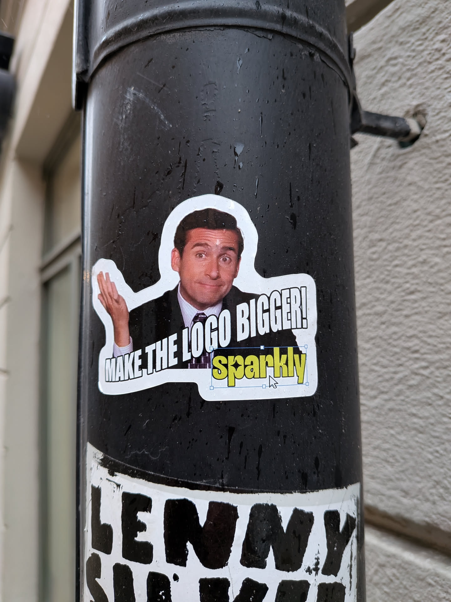 A die-cut sticker featuring a full-color image of Michael Scott (Steve Carell) from The Office, with a slightly bewildered expression and his right hand raised. Below his image, in a speech bubble style, is the text 'MAKE THE LOGO BIGGER!' in white with a black outline. The word 'sparkly' is rendered in yellow with a black outline, positioned below the main text, and includes a small white mouse cursor icon next to it, suggesting a software selection. The entire sticker has a prominent white border and is adhered to a dark, textured pole.