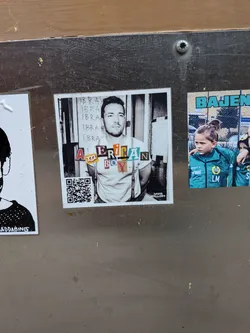 Street sticker The central sticker is square-shaped, showcasing a black and white photographic portrait of a man with a beard, wearing a light-colored t-shirt and suspenders, with his hands clasped behind his back. The background behind him features vertical lines and the repeated word 'IBRAH'. Overlaid on his chest, in a collage style, are the words 'AMERICAN BOY' formed by cut-out letters of various colors, fonts, and textures. In the bottom right corner, small white text reads 'DAVIS MALLORY'. A black and white QR code is present in the bottom left corner.