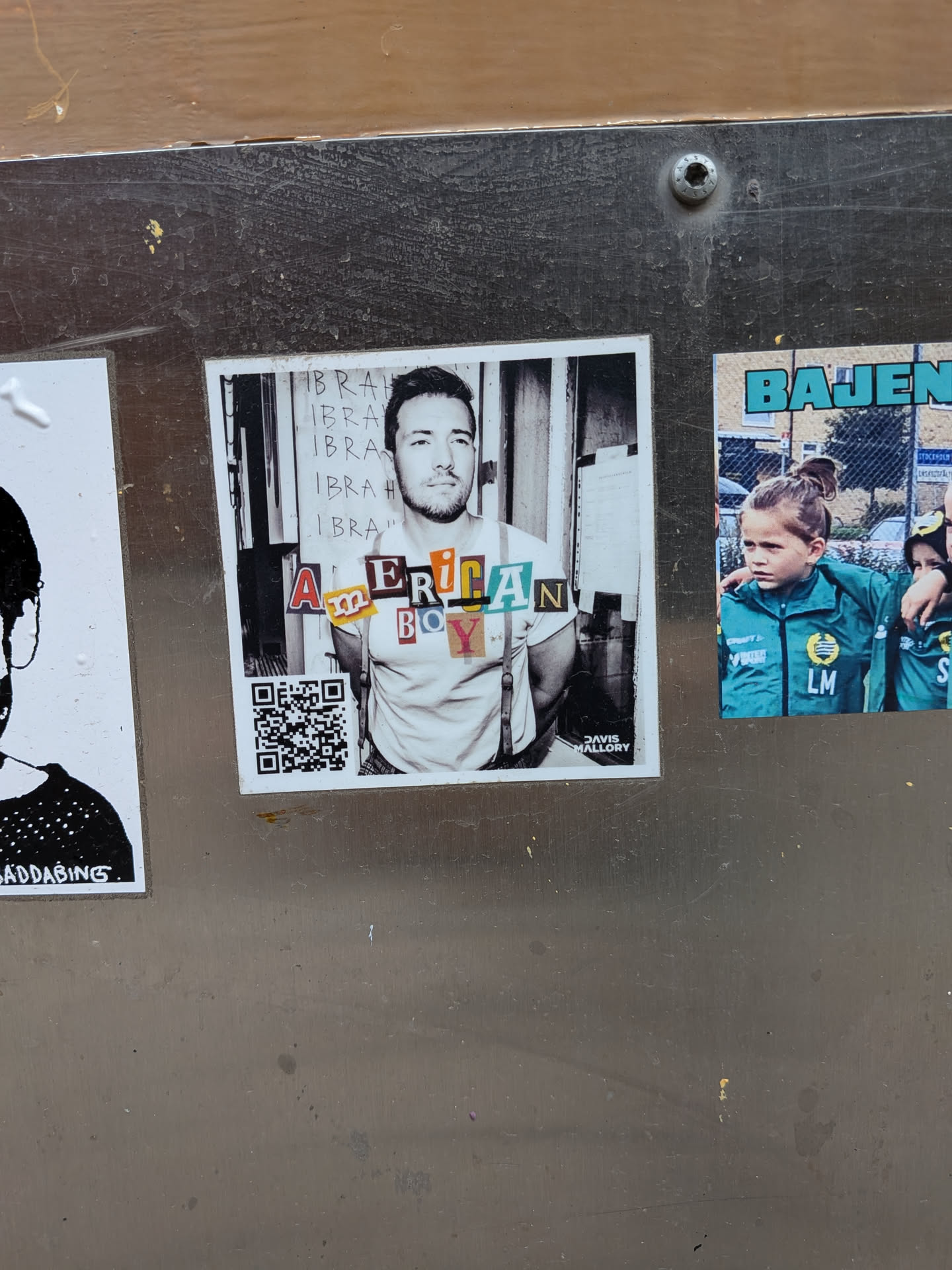 The central sticker is square-shaped, showcasing a black and white photographic portrait of a man with a beard, wearing a light-colored t-shirt and suspenders, with his hands clasped behind his back. The background behind him features vertical lines and the repeated word 'IBRAH'. Overlaid on his chest, in a collage style, are the words 'AMERICAN BOY' formed by cut-out letters of various colors, fonts, and textures. In the bottom right corner, small white text reads 'DAVIS MALLORY'. A black and white QR code is present in the bottom left corner.