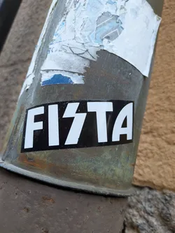 Street sticker A rectangular sticker featuring a solid black background with bold, white, uppercase letters that spell out 'FI7TA'. The middle character '7' is stylized with sharp angles. The sticker is adhered to a weathered, oxidized metal pipe showing signs of rust and green patina, with remnants of torn paper stickers visible above it.