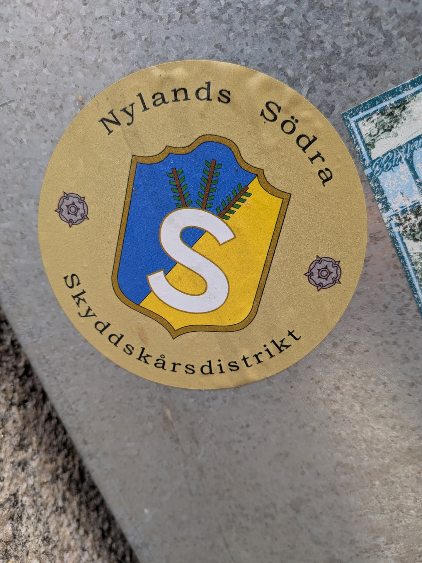 Circular beige sticker featuring a blue and yellow shield with a white letter 'S' and three green spruce twigs. Arched black text reads 'Nylands Södra' above and 'Skyddskårsdistrikt' below, flanked by two small purple roses.