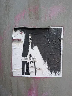 Street sticker The sticker is predominantly white with a large, irregular patch of thick, black paint obscuring the right side and top. A ghostly, black silhouette of a face and upper torso is visible on the white portion, with the black paint appearing to drip and splatter. The text 'RIME OF MNNE' is written in a gothic-style font across the lower center of the white area. The sticker is wrinkled and torn, particularly along the top edge, revealing underlying paper and some pink spray paint on the wall behind. The texture of the black paint is uneven and appears somewhat glossy in places.