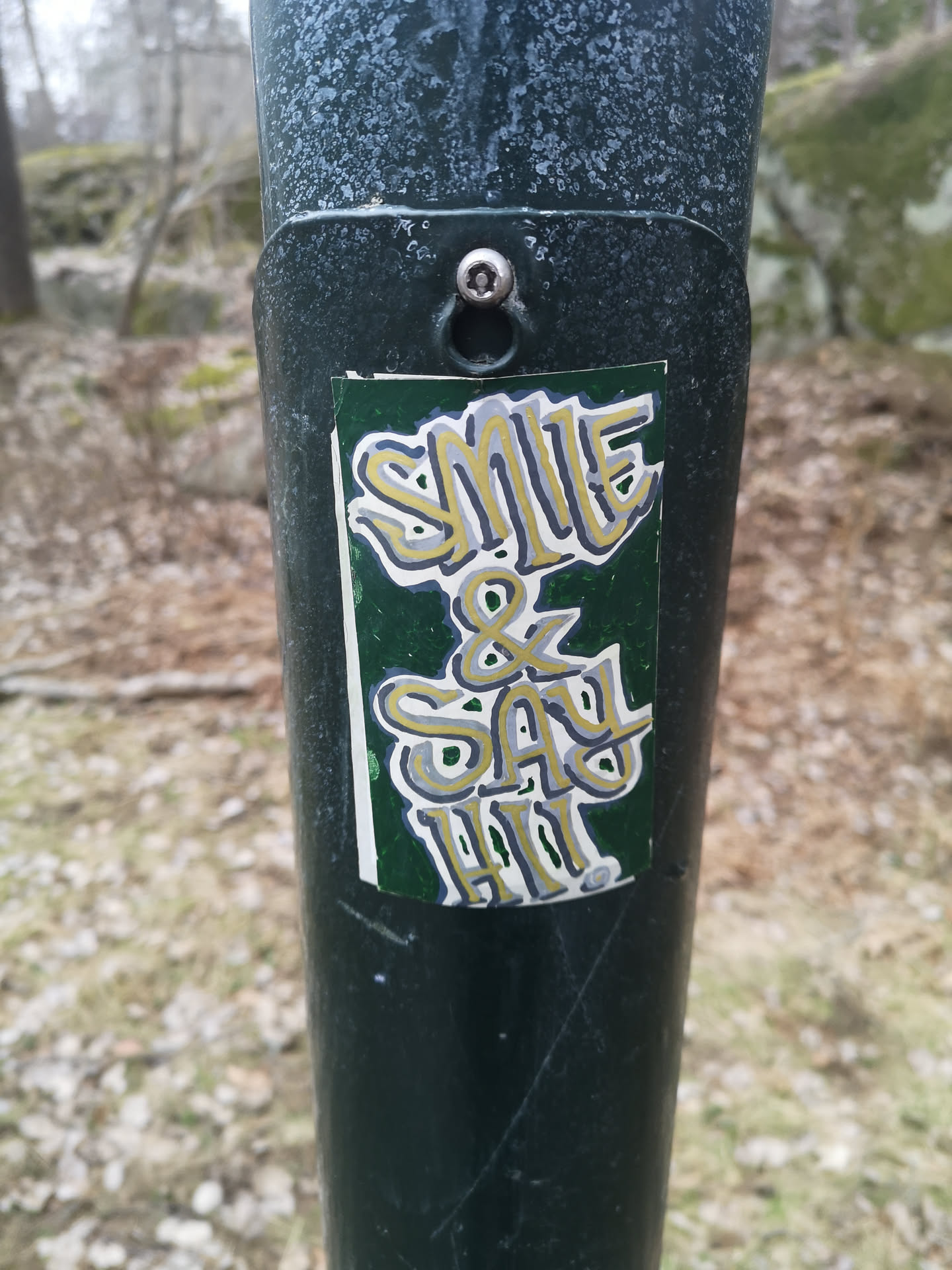 The sticker is a vertically oriented rectangle with a dark green background. It features the hand-lettered text "SMILE & SAY HI!" in a stylized, bold, and somewhat bubbly font. The letters are primarily colored in shades of yellow/gold and grey/silver, outlined with a darker grey or black, and have a white inner highlight or outline, giving them a layered, almost 3D appearance. The overall style suggests a custom, possibly hand-drawn, street art aesthetic. The sticker shows some signs of wear, with slightly peeling edges and minor scuffs, indicating it's a physical sticker applied to a textured, dark green pole.