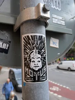 Street sticker The sticker is predominantly black and white. It features a stylized portrait of a person's head, with a sharp, angular design. A sword or similar pointed object is positioned vertically on top of the head. The words 'MOTUS' and 'ATTACK DEFEND' are prominently displayed. The background behind the face has radiating lines, giving it a dynamic and attention-grabbing appearance. The overall style is graphic and somewhat aggressive.