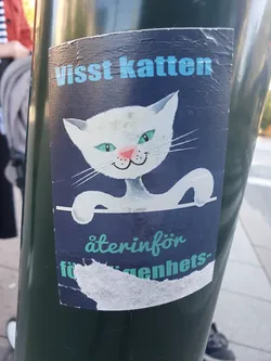 Street sticker A sticker featuring a white cat with teal eyes. The text on the sticker says "Visst Katten" at the top and  "återinför förigenhets-" at the bottom. The sticker is slightly damaged.