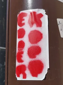 A sticker with red abstract shapes and letters. The letters are slightly visible but hard to read completely. The shapes appear to be dots or circles. The sticker is stuck on a brown pole.