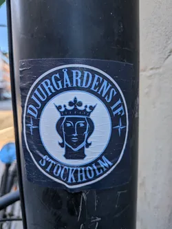 Street sticker The sticker is circular and features a dark blue and light blue color scheme. The outer ring of the circle has text in a light blue, serif font. The top part reads 'DJURGÅRDENS IF', and the bottom part reads 'STOCKHOLM'. Inside the circle, there is a stylized illustration of a woman's head wearing a crown, rendered in dark blue. The background of the inner circle is white, and there are four-pointed star elements on either side of the central illustration, also in light blue. The sticker shows some signs of wear and creasing, indicating it has been applied to a surface.