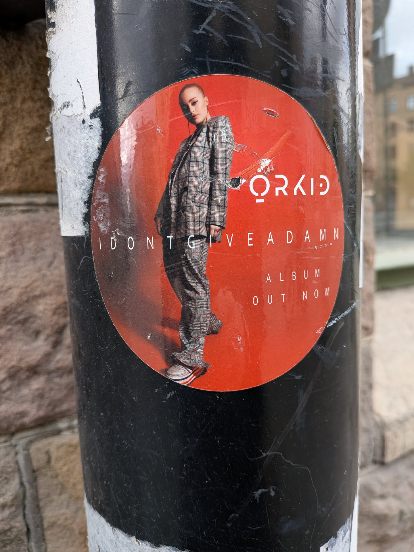A circular promotional sticker featuring a young woman with light skin and hair pulled back, wearing a stylish plaid suit (blazer and trousers). She is depicted in a dynamic pose, leaning slightly. The background of the sticker is a vibrant red-orange. White text on the sticker reads 'ORKID' at the top right, 'I DONT GIVE A DAMN' across the center, and 'ALBUM OUT NOW' below it. The sticker shows signs of wear, with visible creases, minor tears, and a slightly glossy, reflective surface, indicating it's a physical sticker affixed to a black pole.