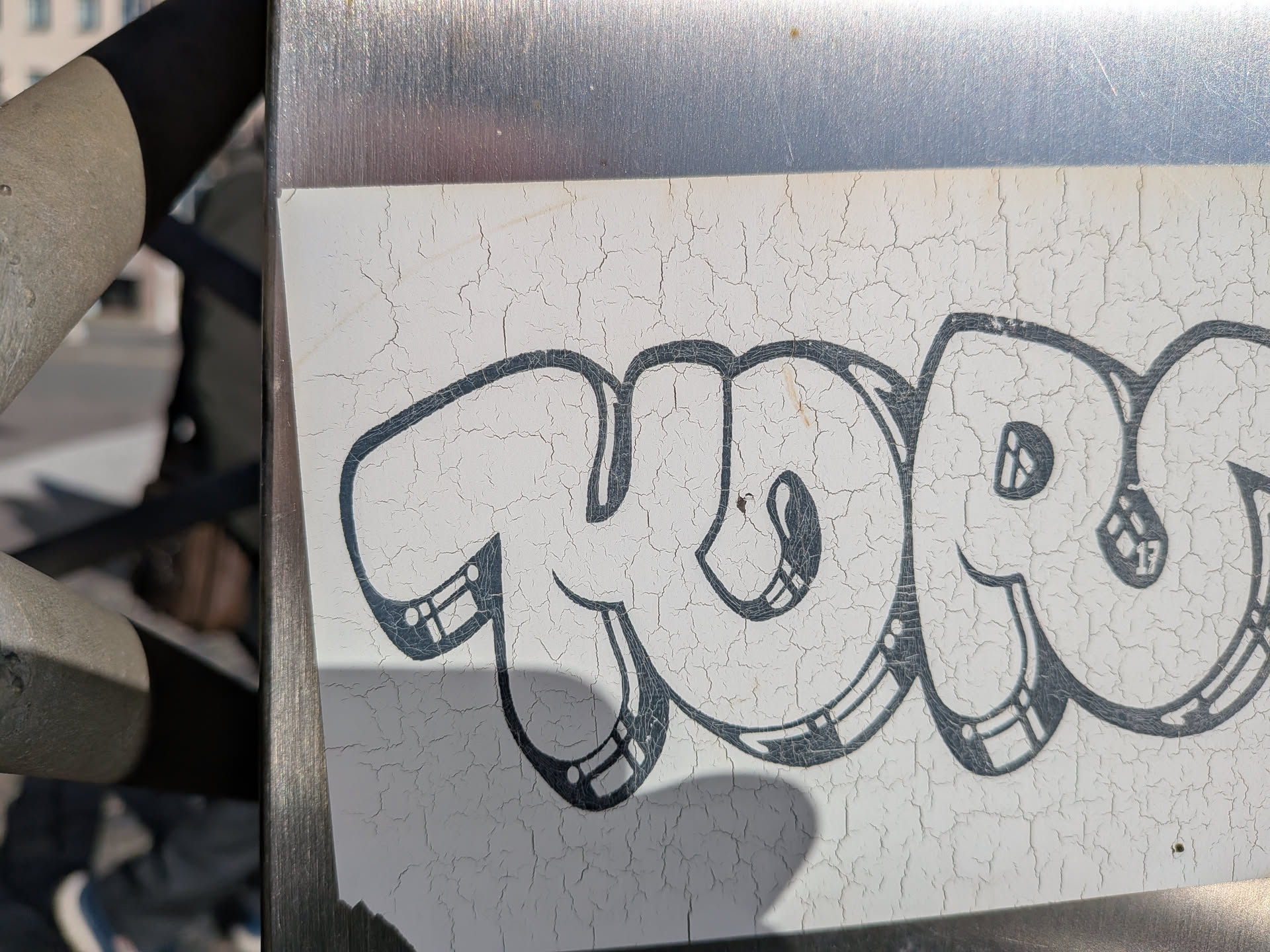 The sticker is white with a distressed, cracked surface. It features bold, black bubble letters that form a word or tag. The letters have thick outlines and some internal detailing, such as parallel lines and small circles, giving them a stylized, graffiti-like appearance. The overall impression is one of age and exposure to the elements.