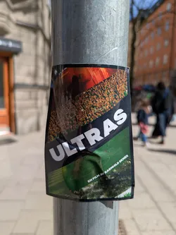 Street sticker This sticker is a poster for the movie "Ultras." The top of the sticker shows a crowd of people in red and orange. The word "ULTRAS" is printed in white letters diagonally across the image. The bottom section shows a view of a soccer field. The sticker is affixed to a pole.