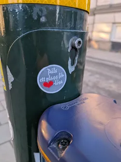 A circular sticker with a light grey background and a thin white border, affixed to a dark green pole. The sticker features white text in a handwritten style. The top line reads 'Däls ett plagg för' and the bottom line reads '-lösa'. Between these two lines, centrally placed, is a vibrant red heart symbol. The sticker shows minor signs of wear, consistent with outdoor placement.