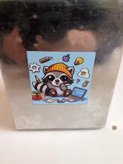 Street sticker A square sticker featuring a charming, cartoon-style raccoon with a light blue background. The raccoon is depicted sitting, wearing a bright yellow beanie, and has a friendly expression. Its fur is shades of grey and white, with black markings around its eyes and striped tail. Surrounding the raccoon are various icons representing technology and creativity: a white gear, a purple Apple logo, a yellow lightbulb, a white cloud with the colorful Microsoft Windows logo, a silver laptop displaying 'Microsoft' on its screen, a paint palette, a pencil, two red cherries, and a question mark. Small colorful items, possibly markers or capsules, are also scattered around. The overall style is cute and vibrant.