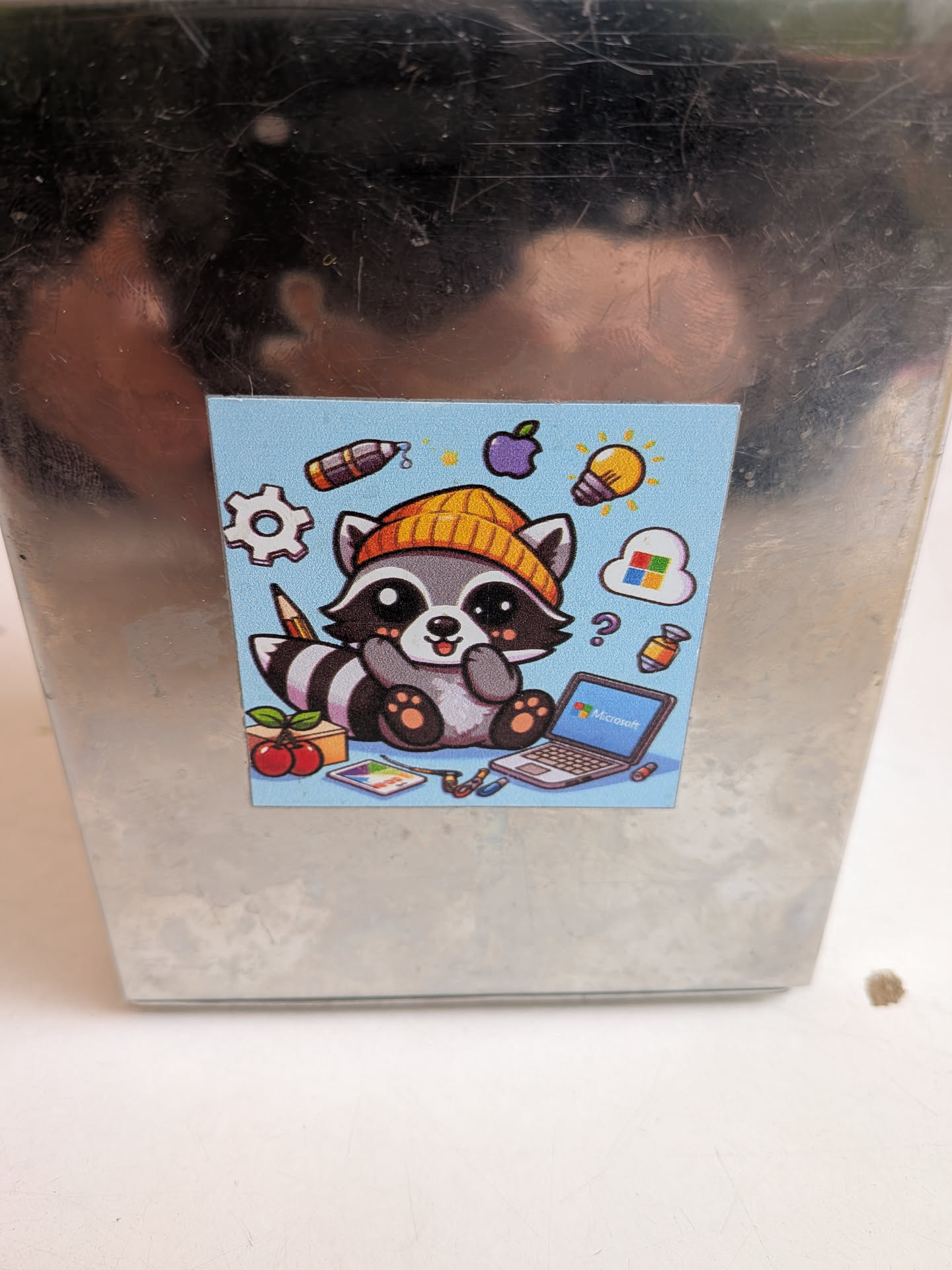 A square sticker featuring a charming, cartoon-style raccoon with a light blue background. The raccoon is depicted sitting, wearing a bright yellow beanie, and has a friendly expression. Its fur is shades of grey and white, with black markings around its eyes and striped tail. Surrounding the raccoon are various icons representing technology and creativity: a white gear, a purple Apple logo, a yellow lightbulb, a white cloud with the colorful Microsoft Windows logo, a silver laptop displaying 'Microsoft' on its screen, a paint palette, a pencil, two red cherries, and a question mark. Small colorful items, possibly markers or capsules, are also scattered around. The overall style is cute and vibrant.