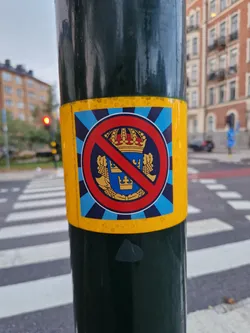 A sticker featuring a red circle-slash symbol over a coat of arms, likely representing opposition to monarchy.  The sticker has a blue and yellow color scheme and is affixed to a pole.