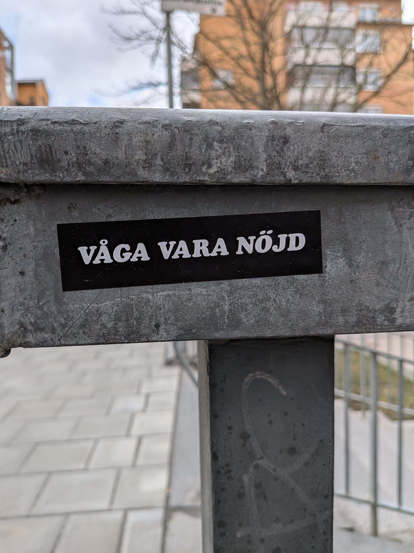 A rectangular black sticker with bold white uppercase text, reading 'VÅGA VARA NÖJD'. The sticker is applied horizontally to a weathered, grey metal beam, showing slight wear and tear around its edges. The text uses a clear, sans-serif font. The overall aesthetic is minimalist and direct.