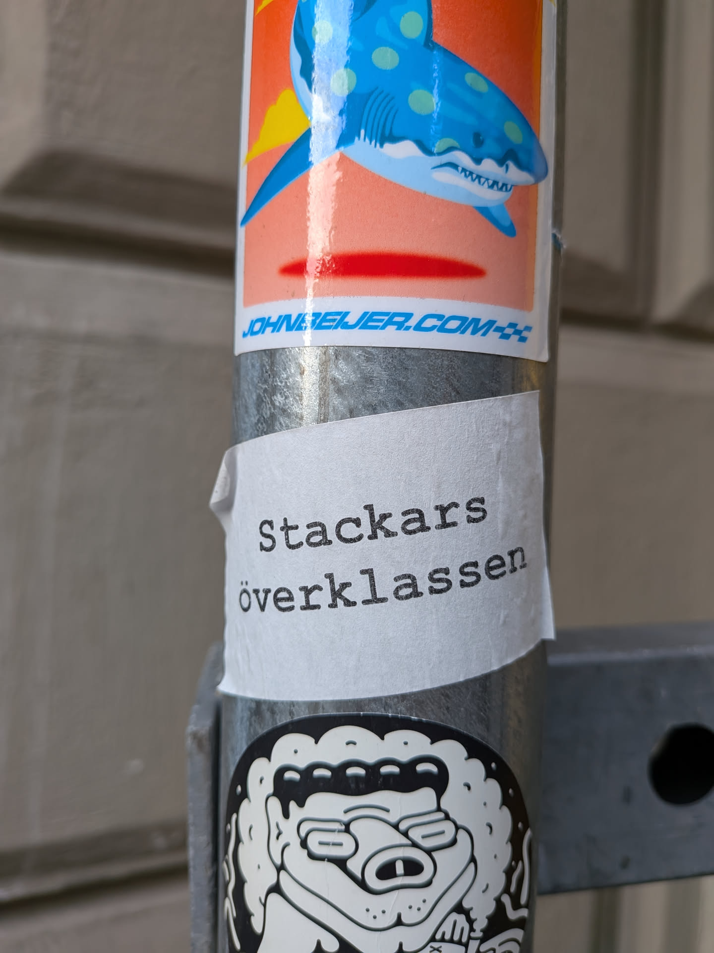 A white sticker with a slightly wrinkled texture, affixed to a metal pole. The sticker has black text printed on it in a serif font. Below it, another black and white sticker with a cartoonish, somewhat menacing creature can be seen.